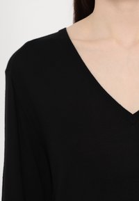 Femme portant un pull noir à col en V, montrant l'encolure et une partie de l'épaule sur un fond uni.