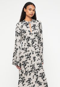 Robe maxi florale en beige avec des motifs floraux noirs, manches longues, décolleté en forme de trou de serrure et coupe ample, fabriquée dans un tissu doux et texturé.
