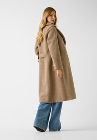 Beiger, strukturierter Mantel mit Schalkragen, doppelreihigem Schnitt und goldenen Knöpfen. Kombiniert mit weitem blauen Jeans.