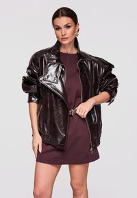 Kobieta ubrana w błyszczącą, ciemnobrązową, oversize'ową skórzaną kurtkę na krótkiej bordowej sukience z paskiem, stojąca na tle jednolitego, jasnego tła.