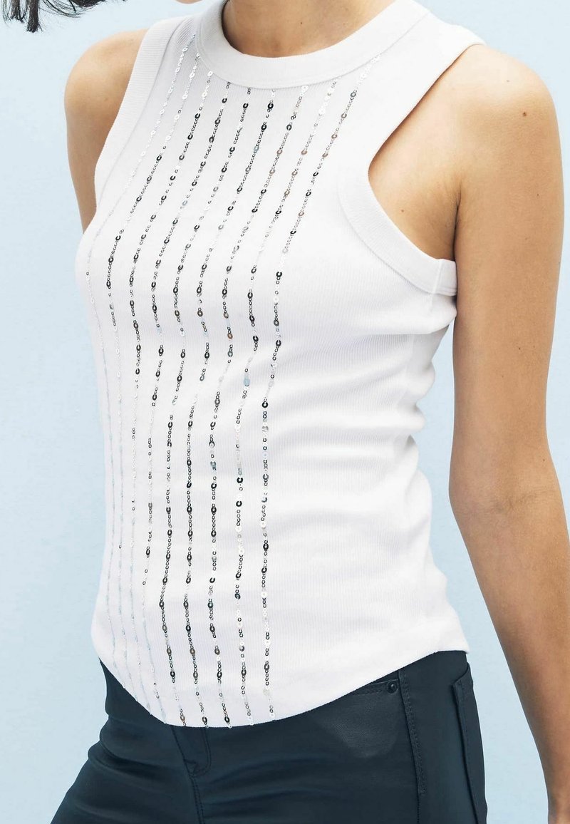 Haut sans manches blanc avec une texture côtelée, présentant des lignes verticales de sequins argentés sur le devant. Encolure ronde, coupe ajustée.