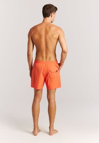 Pantaloncini da bagno arancioni vivaci con una cintura elastica in vita e tasca posteriore, caratterizzati da una texture liscia e una lunghezza a metà coscia.