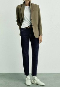 Blazer beige ajustado sobre una camiseta blanca, combinado con jeans azul oscuro y zapatillas blancas. Las características incluyen un bolsillo en el pecho y un cinturón.