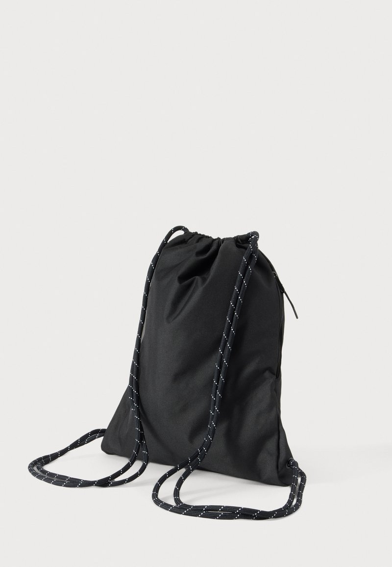 Black drawstring backpack with white dotted cords, simple soft fabric design, standing on a plain light background.