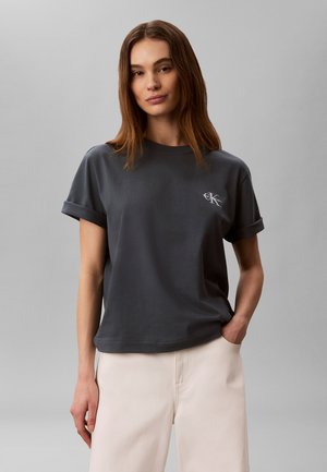 Femme aux longs cheveux bruns portant un t-shirt gris foncé à manches retroussées avec un logo blanc et un pantalon beige clair, debout devant un fond uni.
