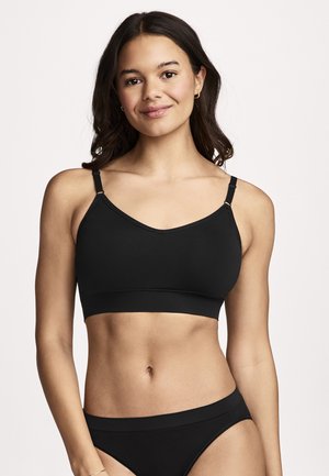 Junge Frau mit langen dunklen Haaren trägt schwarzen BH mit verstellbaren Trägern und passende schwarze Unterwäsche, lächelt leicht und schaut nach vorne.