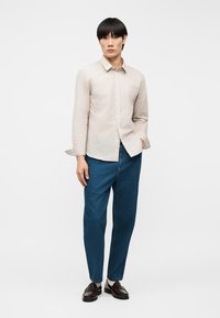 Νεαρός άνδρας στέκεται φορώντας μια μπεζ πουκάμισο με κουμπιά, μπλε τζιν, μπεζ κάλτσες και σκούρα καφέ δερμάτινα loafers μπροστά σε λευκό απλό φόντο.