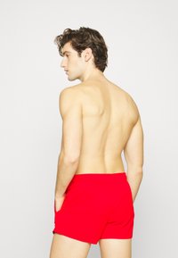 Rote Badeshorts mit elastischem Bund und Seitentaschen, von hinten gegen einen schlichten weißen Hintergrund gezeigt.