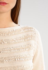 Maglione cream lavorato a maglia con un motivo testurizzato caratterizzato da strisce orizzontali e dettagli frangianti sulla superficie.