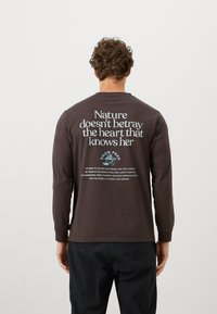 Long-sleeve brown t-shirt made of cotton with white and blue text on the back, featuring a nature-inspired quote and a small graphic logo.