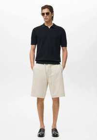 Polo negro de manga corta con punto texturizado, combinado con shorts beige claros y mocasines negros. Gafas de sol complementan el look.