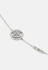 Silberarmband mit einem runden Anhänger, der ein Logo und Kristallakzente zeigt, befestigt an einer eleganten Kette mit dem Markennamen "GUESS."