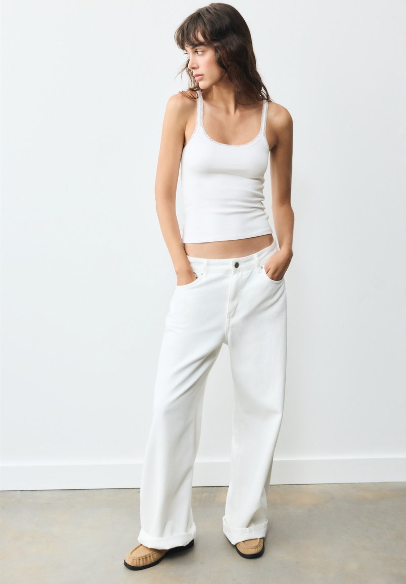 Femme debout, les mains dans les poches, portant un débardeur blanc, un pantalon large blanc et des mocassins beiges sur un fond uni.