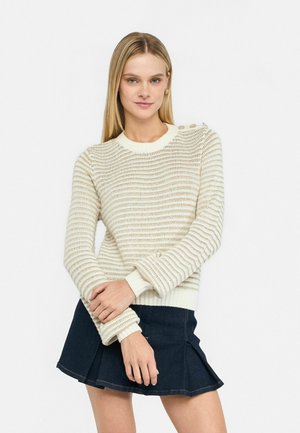 Maglione lavorato a maglia di colore crema con righe orizzontali, dotato di scollatura tonda, polsini a coste e dettagli con bottoni sulla spalla. Abbinato a una gonna di jeans.