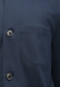 Nærbilde av mørk marineblått stoff med to svarte firehullsknapper og en sydd lomme på et plagg.