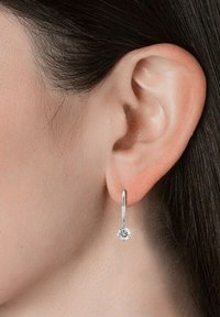 Orecchino a cerchio in argento con design a goccia decorato da un accento di pietra preziosa trasparente. L'orecchino pende appena sotto il lobo dell'orecchio, riflettendo la luce.