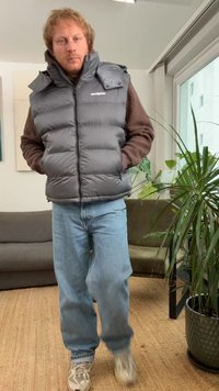 Graue Steppweste mit hohem Kragen, braunen Fleecepuffärmeln und Reißverschlusstaschen, kombiniert mit hellblauen Jeans und Turnschuhen. Innenraum mit Pflanzen.