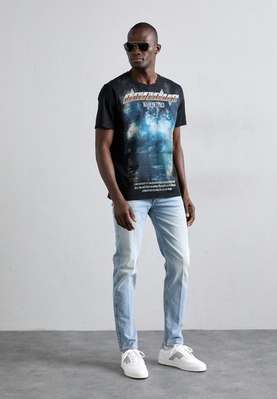 Musta graafinen t-paita, jossa on värikäs printti ja teksti, vaaleansiniset farkut ja valkoiset lenkkarit, joissa on harmaita yksityiskohtia. Aurinkolasit täydentävät asun.