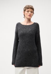 Temno siva oversize jopica s krožnim ovratnikom, dolgimi rokavi in teksturnim pletenim vzorcem, v kombinaciji z belimi hlačami.