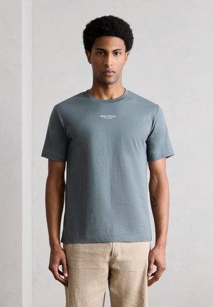 Άντρας που φοράει γκρι μπλουζάκι Marc O’Polo και μπεζ παντελόνι, στέκεται μπροστά σε απλό ανοιχτόχρωμο φόντο, κοιτάζοντας προς τα εμπρός.