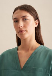 Top verde a scollo a V realizzato in tessuto morbido, con taschino sul petto e maniche corte. Texture liscia e vestibilità rilassata.