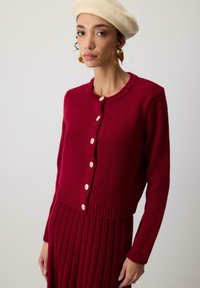 Czerwony kardigan z dzianiny, z długimi rękawami, złotymi guzikami i prążkowanym wykończeniem, zestawiony z dopasowaną spódnicą o prążkowanej fakturze. Materiał z mieszanki wełny.