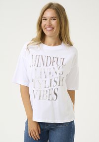 T-shirt blanc à manches courtes avec un texte argenté métallisé indiquant "VIE CONSCIENTE, VIBES STYLÉES", associé à un jean en denim bleu.