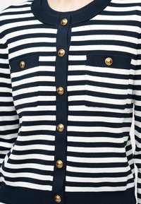 Ναυτικό μπλε και λευκή ριγέ ζακέτα με χρυσά στρογγυλά κουμπιά και δύο εμπρόσθιες τσέπες, φορεμένη από άτομο από τον λαιμό μέχρι τη μέση.