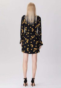 Femme aux longs cheveux blonds portant une robe noire avec des motifs de fruits jaunes et des chaussures à talons hauts avec bride de cheville noires, se tenant de dos.