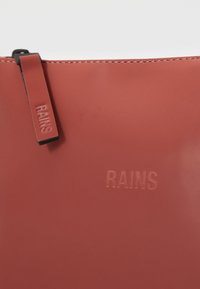 Borsa a pochette rossa lucida realizzata in materiale impermeabile, con logo 'RAINS' impresso e una cerniera nera. Texture liscia su tutta la superficie.