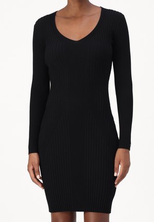 Strakke zwarte geribbelde gebreide jurk met lange mouwen en een V-hals, gedragen door een staande persoon met ontspannen handen langs het lichaam.