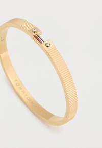 Βραχιόλι σε χρυσό τόνο με ραβδώσεις, διακοσμημένο με δύο διάφανα κρύσταλλα και μια μικρή λεπτομέρεια με κόκκινες, λευκές και μπλε ρίγες κοντά στον κούμπωμα.