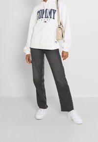 Persona vistiendo una sudadera blanca de Tommy Jeans, jeans negros, zapatillas blancas y llevando un bolso de hombro beige, de pie contra un fondo blanco.