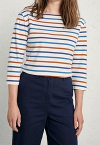 Gestreepte lange mouwen top in het wit, met horizontale strepen in blauw, oranje en marineblauw. Ronde halslijn en een ontspannen pasvorm. Gekleed met marineblauwe broeken.