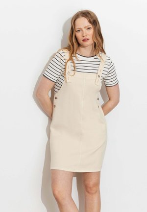 Jeune femme aux longs cheveux portant une robe salopette beige par-dessus un t-shirt à manches courtes rayé blanc et noir, debout devant un fond blanc.