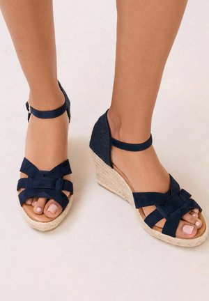 Feet wearing navy blue wedge sandals with ankle straps and woven espadrille-style heels on a light background.