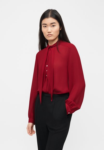 Femme portant un chemisier rouge à manches longues avec un nœud au col et un pantalon noir taille haute, debout devant un fond clair uni.