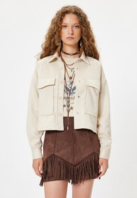 Chaqueta corta color crema con dos bolsillos delanteros, combinada con una falda de ante marrón con detalles de flecos y una camiseta gráfica debajo.