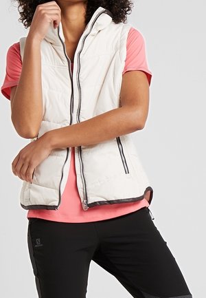 Personne portant un gilet matelassé blanc par-dessus un t-shirt rose à manches courtes et un pantalon noir, debout avec un bras croisé et une main touchant le menton.