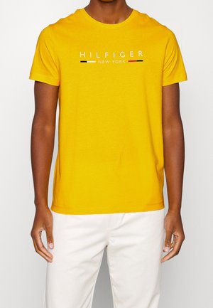 Mężczyzna w żółtym krótkim rękawem z napisem "HILFIGER NEW YORK" i kolorowymi paskami, w białych spodniach, na białym tle.