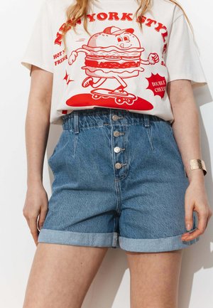 Femme portant un short en jean retroussé taille haute et un T-shirt blanc avec un motif rouge représentant un cheeseburger en train de faire du skateboard.
