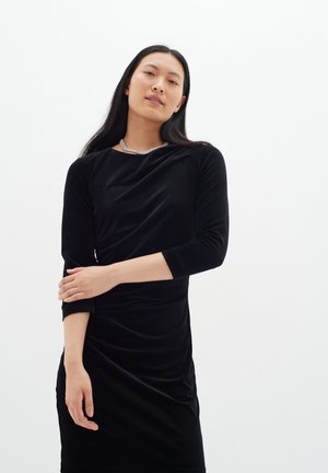 Svart sammetklänning med trekvartslånga ärmar, draperad design och en slät yta, accessoriserad med ett silverhalsband.