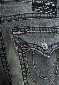 Ciemnoszara kieszeń z denimu z klapą i metalowym guzikiem, kontrastujące beżowe szwy oraz czarna metka z białym tekstem i wzorem misia grającego na gitarze.