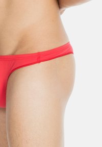 Rote Stoff-Bikini-Unterwäsche mit schmalem Bund, glatter Textur und minimaler Bedeckung, die eine Seitenansicht des Körpers zeigt.