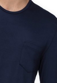 Mężczyzna w jednolitym ciemnoniebieskim T-shircie z okrągłym dekoltem i małą kieszonką na lewej stronie.