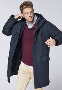 Navy blue long coat with a hood, front zipper, and side pockets, worn over a burgundy sweater and striped collared shirt. Smooth texture.