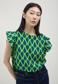 Donna con capelli lisci e scuri, che indossa una maglietta verde con motivi geometrici blu e neri e jeans blu scuro, in piedi davanti a uno sfondo semplice.