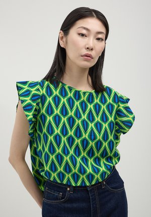 Donna con capelli lisci e scuri, che indossa una maglietta verde con motivi geometrici blu e neri e jeans blu scuro, in piedi davanti a uno sfondo semplice.