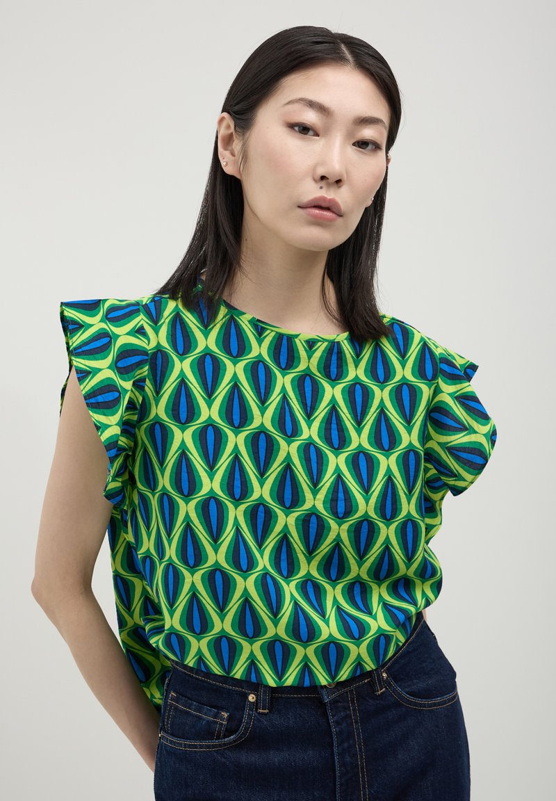 Donna con capelli lisci e scuri, che indossa una maglietta verde con motivi geometrici blu e neri e jeans blu scuro, in piedi davanti a uno sfondo semplice.