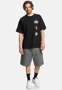 Černé oversized tričko s potiskem log, spárované se šedými džínovými šortkami. Model má na sobě černé ponožky a bílé tenisky.